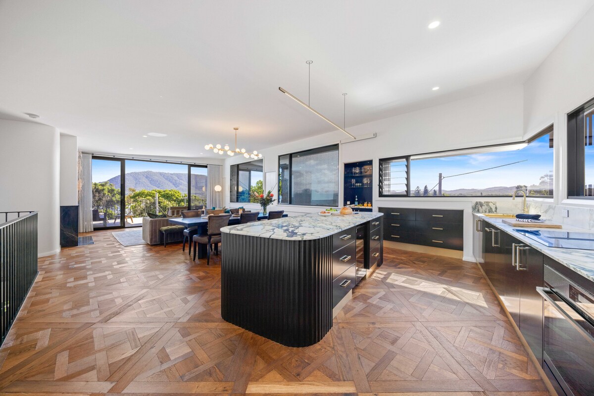 An open-plan kitchen and dining area is highlighted by a large marble island with sleek black cabinetry. Natural light floods the space from expansive windows, revealing surrounding greenery and mountain views, while herringbone-patterned wooden flooring adds warmth to the modern design.