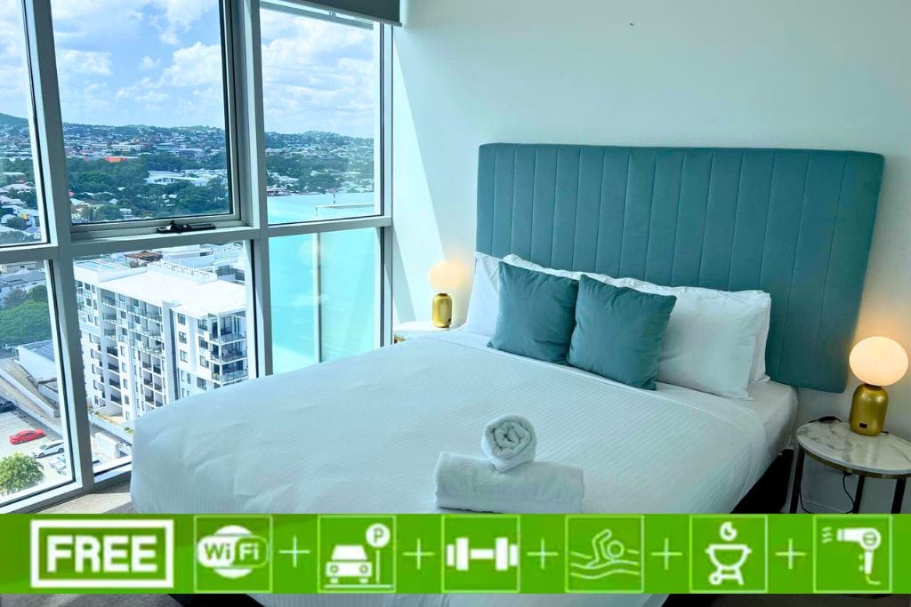 A spacious bedroom features a large bed dressed in fresh linens. Two decorative pillows enhance the clean aesthetic. Views extend from the window, revealing the cityscape beyond. A modern bedside table supports a soft-glowing lamp, contributing to the room's bright and airy feel.