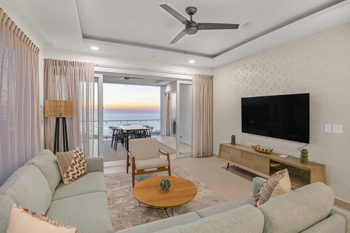 The living area features a light-colored sofa paired with an armchair, arranged around a wooden coffee table. Large glass doors offer ocean views during sunset, while soft drapes frame the windows. A wall-mounted television and minimalistic decor enhance the space's modern design.