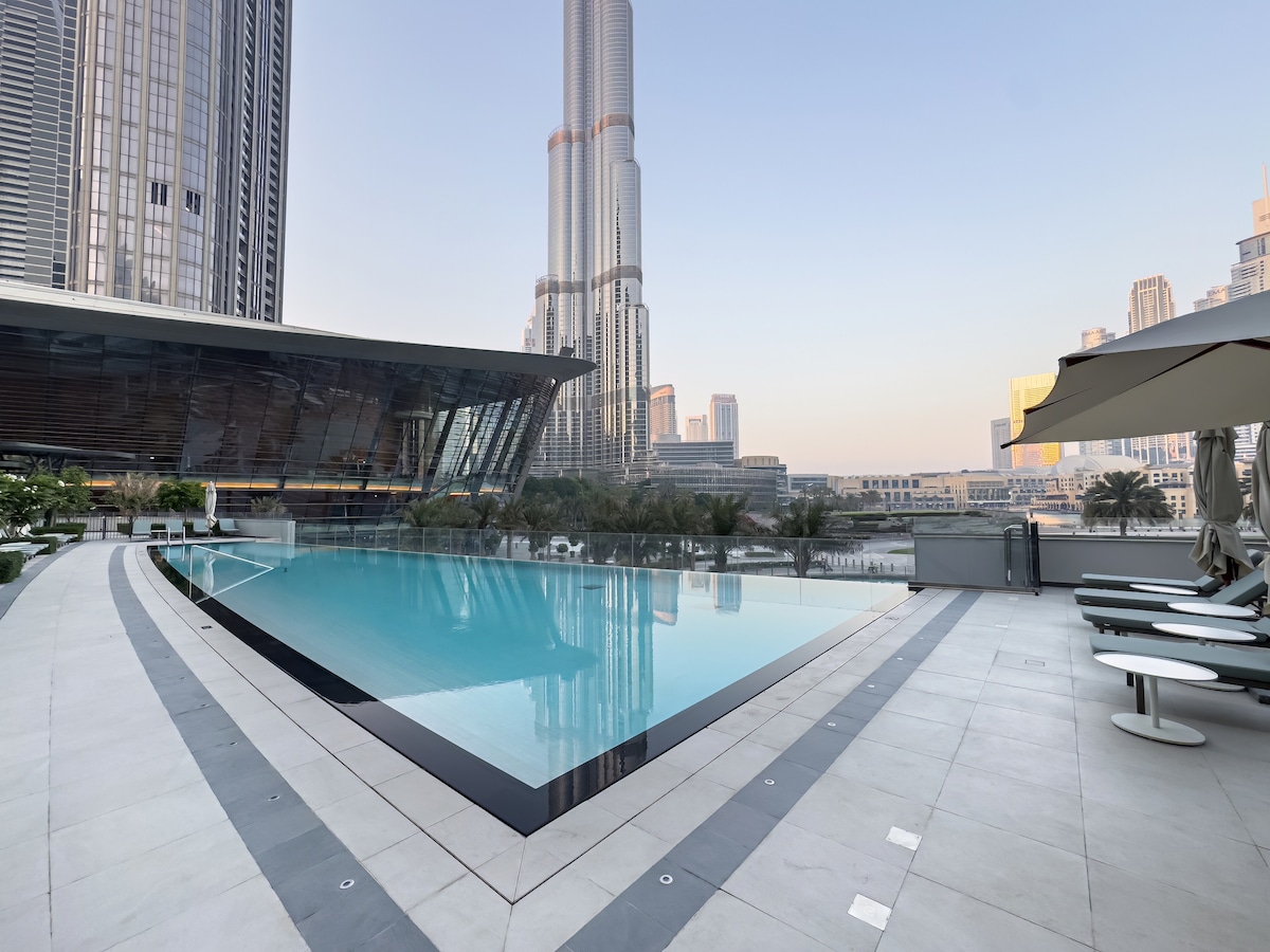 An infinity pool is presented in the foreground, featuring clear water that blends seamlessly with the skyline. The Burj Khalifa stands majestically in the background, while lounge chairs are arranged around the pool's edges. Sun umbrellas provide shaded seating areas.