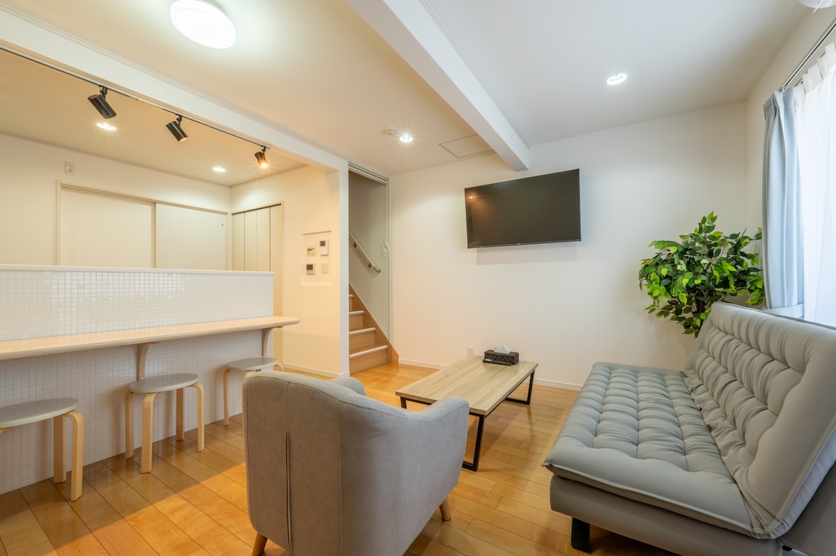 A spacious living area features a comfortable gray sofa with tufted upholstery, positioned beside a wooden coffee table. A flat-screen television is mounted on the wall, while a small dining area with two stools is visible in the background. Natural light enters through a window with light curtains.