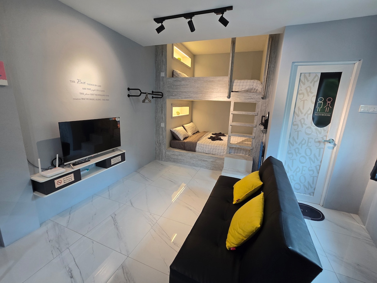 A well-appointed studio features a modern design with light-colored walls and glossy tile flooring. Bunk beds are positioned against one wall, complemented by a sleek sofa adorned with yellow cushions. A flat-screen TV is mounted on the wall, enhancing the functional space.