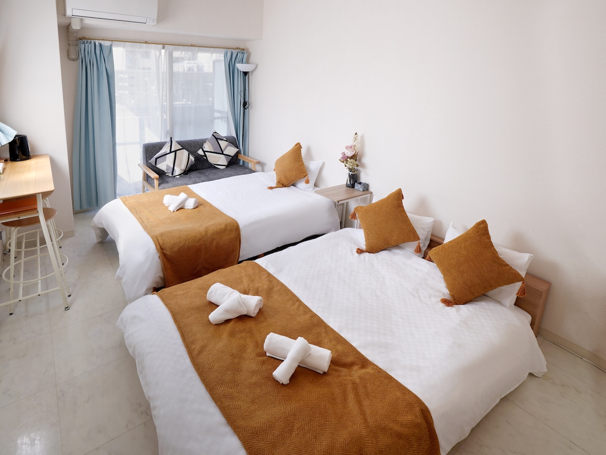 The room features two spacious beds with warm-colored throws and decorative pillows. A window with light blue curtains allows natural light to fill the space. A seating area with cushions is situated near the window, accompanied by a wooden table and bar stools.