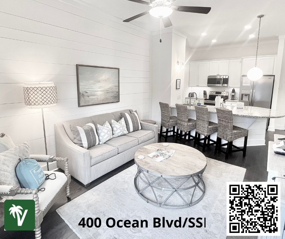 A bright living area features a light-colored sofa adorned with decorative pillows. Soft sunlight illuminates the space, highlighting a round coffee table made of natural wood. In the background, a modern kitchen with bar seating is visible, contributing to a welcoming open-concept design.