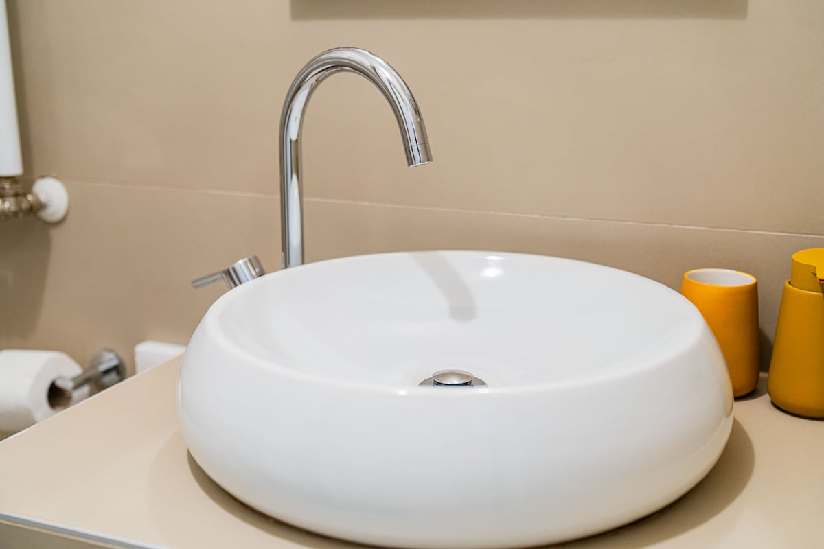 A modern bathroom sink is featured, showcasing a sleek, round basin in white. A tall, polished chrome faucet arcs above the sink. Two yellow cups are positioned nearby, providing a pop of color against the neutral-toned counter and walls.