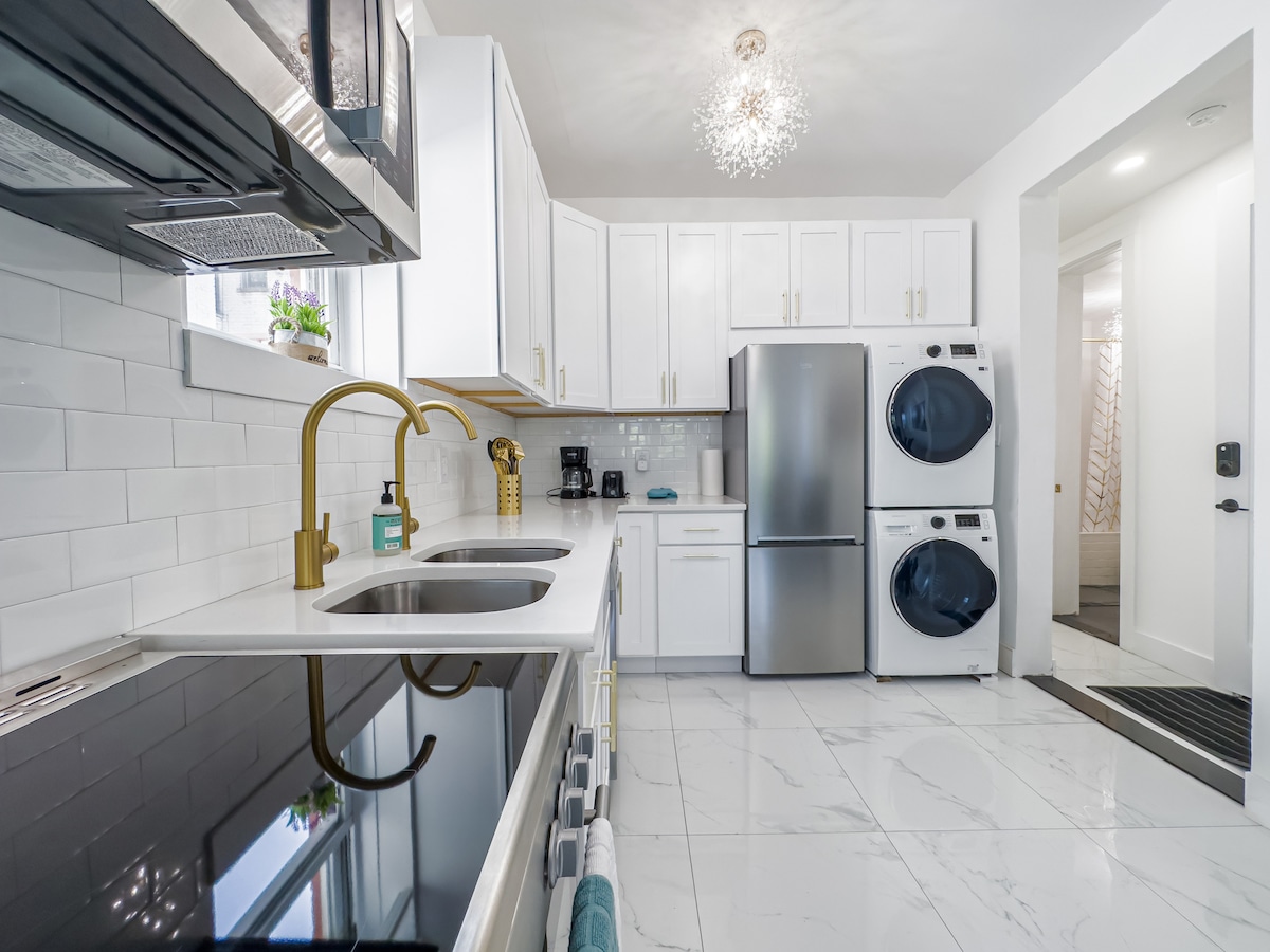 A modern kitchen features sleek white cabinets with gold accents and a white countertop. A double sink is positioned below a window, while a stainless steel fridge and washer-dryer stand against the wall. Natural light enhances the clean, spacious layout.