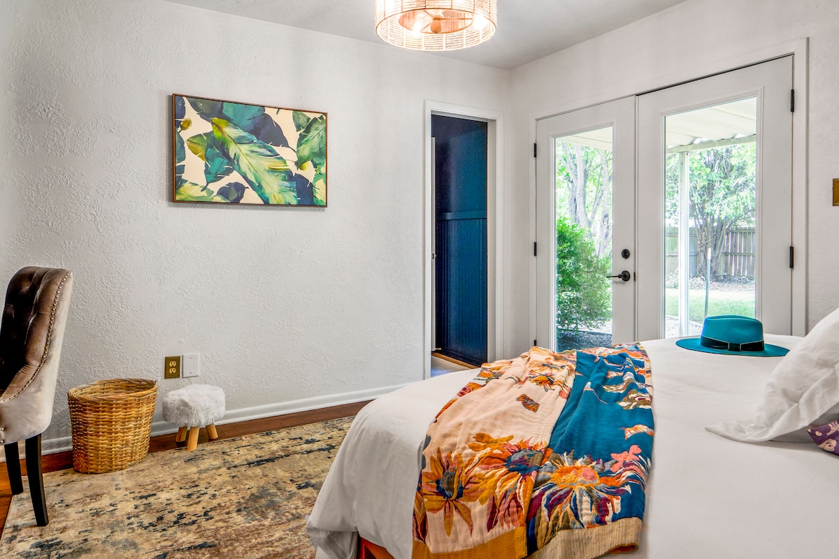 A bright bedroom is featured with large glass doors that open to a garden area. A spacious bed is adorned with a colorful blanket, complemented by a textured rug. A stylish chair and a decorative wall art piece add visual interest to the room.