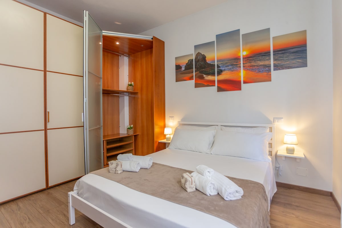 The bedroom features a spacious bed dressed in light bedding, accompanied by neatly arranged towels. A large wardrobe stands beside the bed, with shelves and a sliding door. The wall is adorned with a sunset-themed artwork, enhancing the serene atmosphere.
