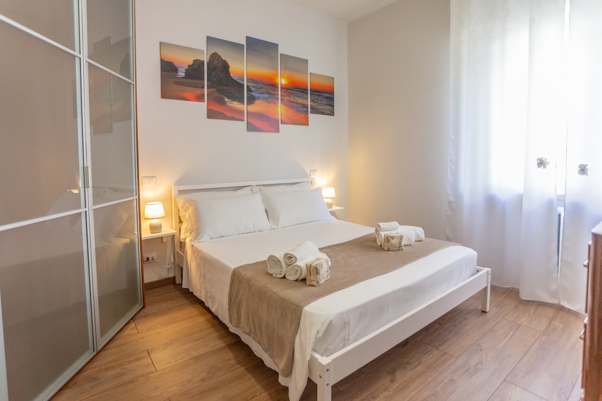 A spacious bedroom features a large bed with white linens and a neutral-colored bedspread. Two bedside lamps provide soft lighting. A decorative wall featuring sunset images complements the room. Natural light filters through sheer curtains, creating a bright and airy atmosphere.