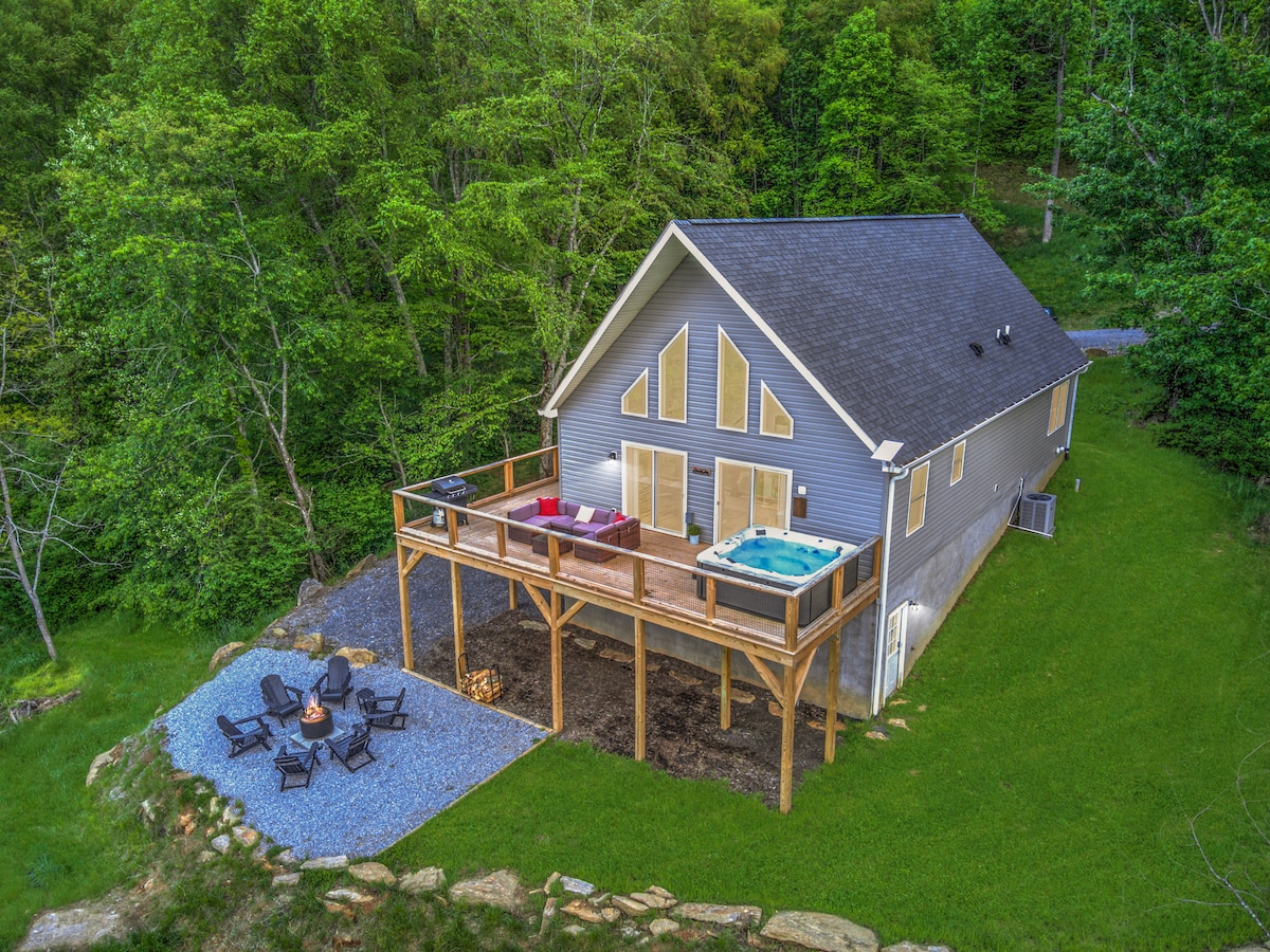 An elevated view showcases the modern farmhouse nestled among green trees. A spacious deck features a hot tub and cozy seating around a stone fire pit, positioned on a gravel area, enhancing the natural surroundings.
