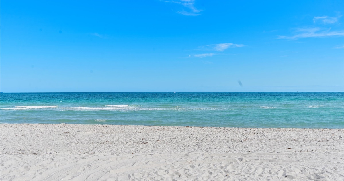 A serene beach scene features soft white sand transitioning into calm turquoise waters. The horizon is visible under a clear blue sky, with gentle waves lapping at the shore. This tranquil setting invites relaxation and enjoyment of the coastal environment.