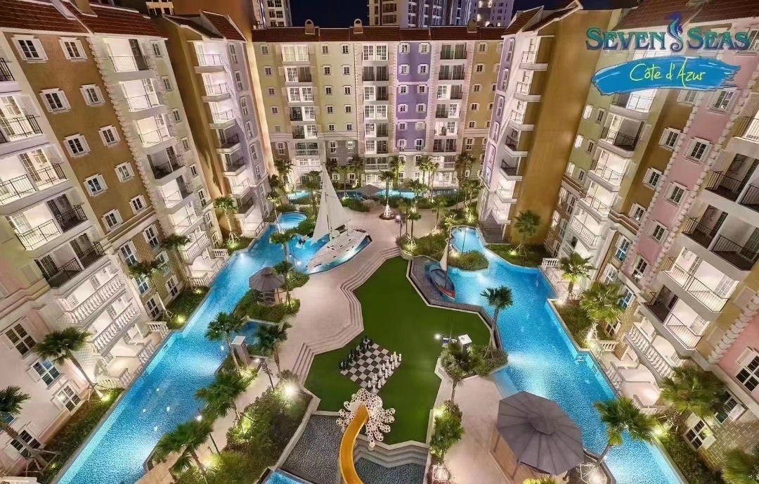 The image displays a resort-like complex featuring multiple lagoon-style swimming pools surrounded by tropical landscaping. Lounge areas with umbrellas are visible, along with manicured green lawns. The building's architecture includes several colorful facades, creating an inviting atmosphere within the bustling community.