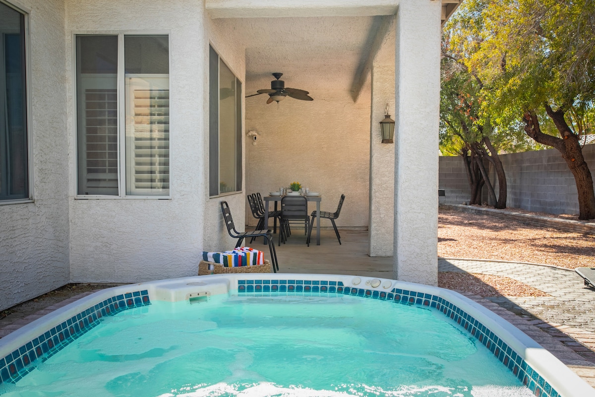 A private spa is situated next to a covered patio area, equipped with a dining table and chairs. A ceiling fan is visible overhead, while surrounding landscaping features mature trees, providing shade and privacy.