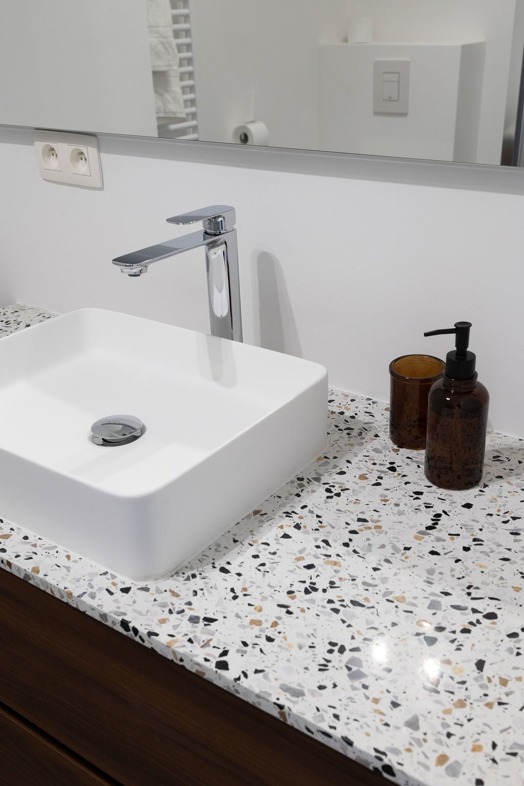 A modern bathroom sink is presented on a patterned terrazzo countertop. The sleek, square sink sits above a wooden cabinet, accompanied by a dark glass soap dispenser and a matching holder. A large mirror reflects the contemporary design.
