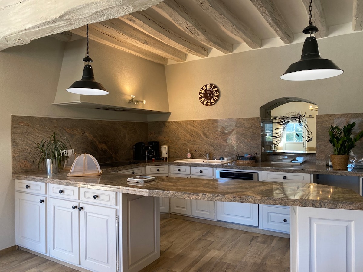 The kitchen features a large central island with a stone countertop, surrounded by ample white cabinetry. Two pendant lights illuminate the space, while a wall clock adds a decorative touch. A coffee maker and plants are included, providing functional and aesthetic elements.