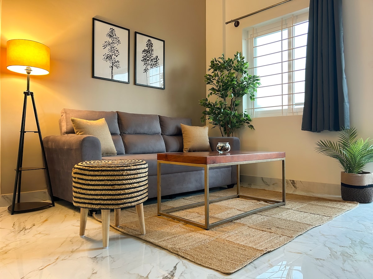 A modern living area is featured with a comfortable grey sofa, accompanied by a decorative stool. Natural light enters through a large window, accented by dark curtains. A coffee table rests on a woven rug, and greenery adds a refreshing touch to the space.
