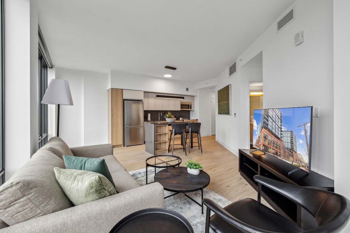 A bright living space features a comfortable sofa with accent pillows, paired with sleek black chairs. A wooden dining table is visible in the background, along with modern kitchen appliances and cabinetry. Large windows allow natural light to fill the area.