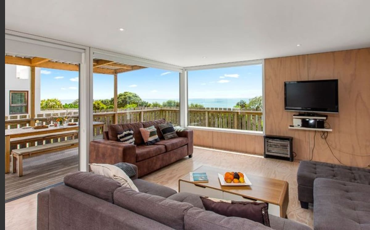 The open-plan living area features large glass doors that provide access to stunning coastal views. Comfortable seating arrangements are present, including dark sofas, with a coffee table displaying fruit. A television is mounted on the wall, enhancing the modern decor.