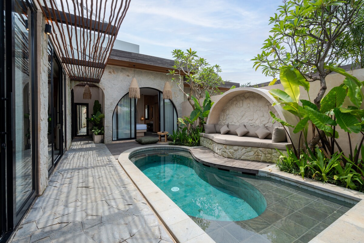 An outdoor area features a serene swimming pool surrounded by lush greenery. A cozy seating area with smooth cushions is positioned under a curved alcove, offering a shaded space to relax. Light filters through the overhead wooden slats, creating a peaceful ambiance.