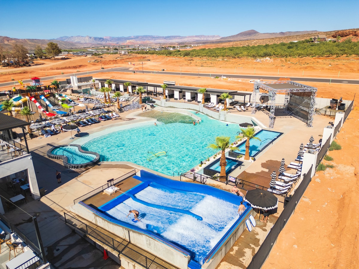 An expansive outdoor water park area is shown, featuring a large swimming pool surrounded by lounge chairs. A water slide is visible alongside a FlowRider surf setup, with palm trees adding greenery to the landscape. The scenic backdrop includes arid hills.