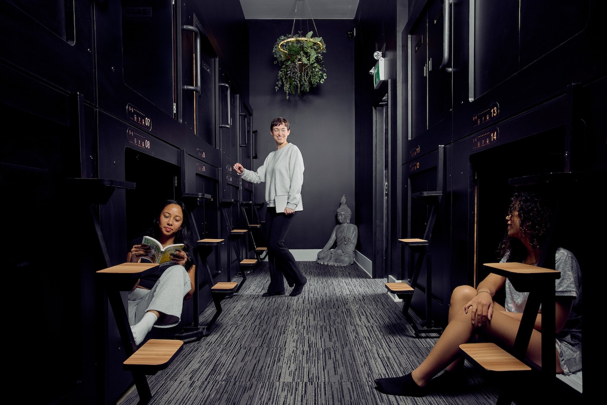 A corridor with individual capsule beds is presented, featuring wooden seating areas with portable tables. Soft lighting highlights the dark walls, while a plant hangs from above. A serene statue adds a calming element to the space, suggesting a modern and relaxed environment.