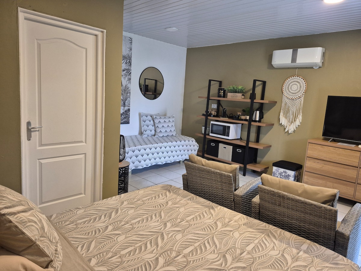 The studio features a comfortable sleeping area with a patterned bedspread. A small seating area with two textured chairs is situated opposite a TV unit and a microwave. A round mirror reflects light, enhancing the space, while an air conditioning unit is mounted on the wall.