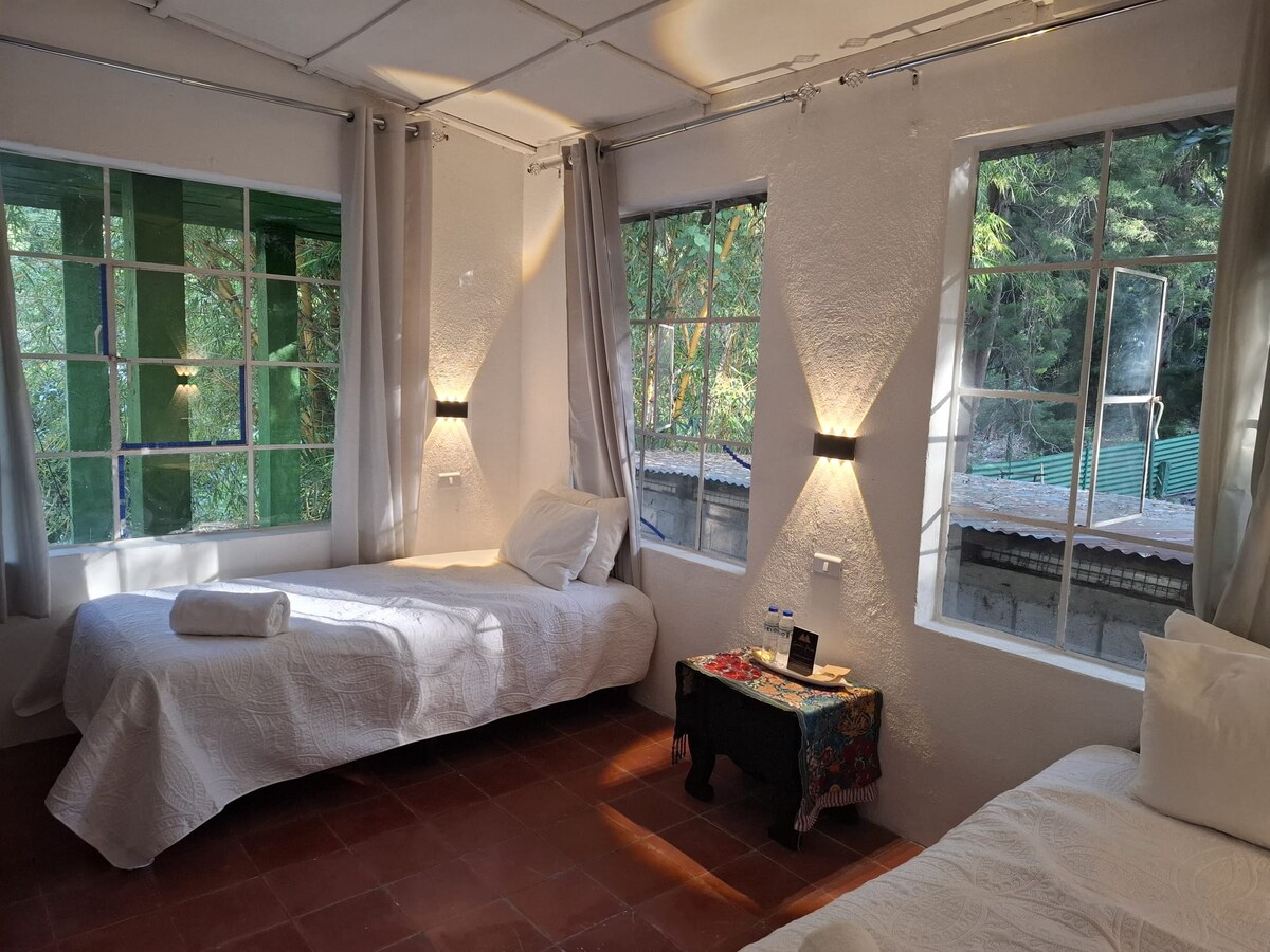 A bright sleeping area is punctuated by two spacious beds, surrounded by large windows that allow natural light to flood in. Soft curtains frame the windows, providing a sense of openness. A small wooden table with decorative cloth offers a touch of local charm.