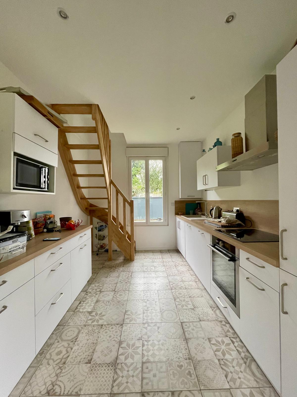 The kitchen features modern cabinetry with an array of appliances, including a microwave and oven. A central window allows natural light to filter in, while a wooden staircase leads to the upper level. Decorative tiled flooring adds a unique touch to the space.