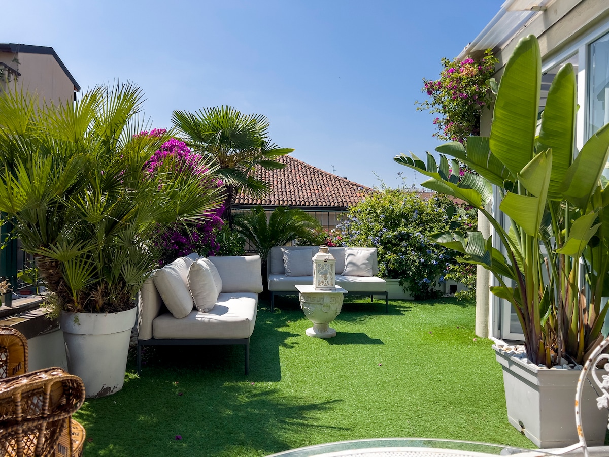 A spacious terrace is adorned with various potted plants and vibrant flowers. Comfortable seating arrangements are provided, along with a decorative stone feature. The area is surrounded by greenery, creating a tranquil outdoor setting with clear blue skies overhead.