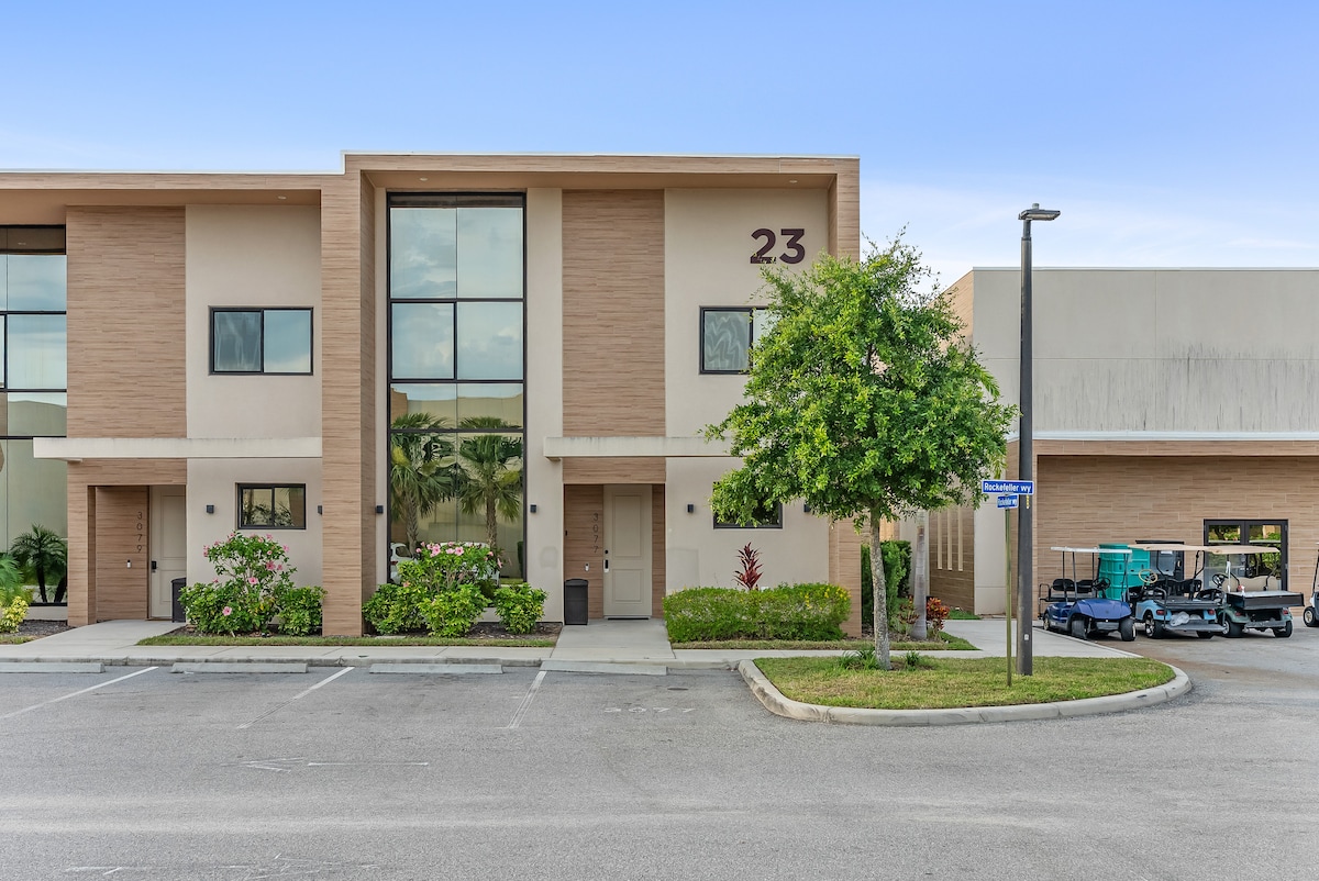 The facade of a townhouse is shown, featuring a large central entrance flanked by symmetrical windows. The area includes trimmed shrubbery and small trees, while a parking lot is visible in the foreground, containing several empty spaces.