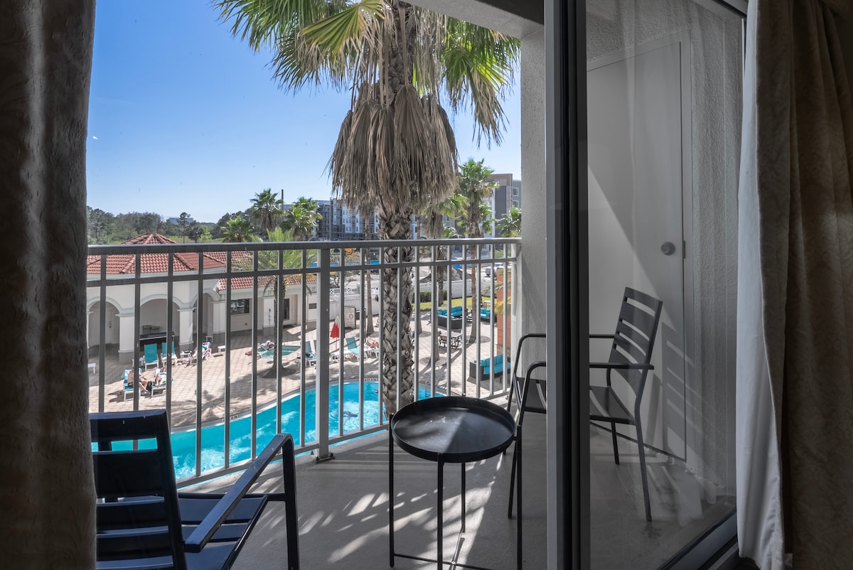 A private balcony is shown featuring outdoor furniture, including a small round table and chairs. Lush palm trees are visible, framing the view of a pool area where guests can be seen enjoying the sunshine and water.