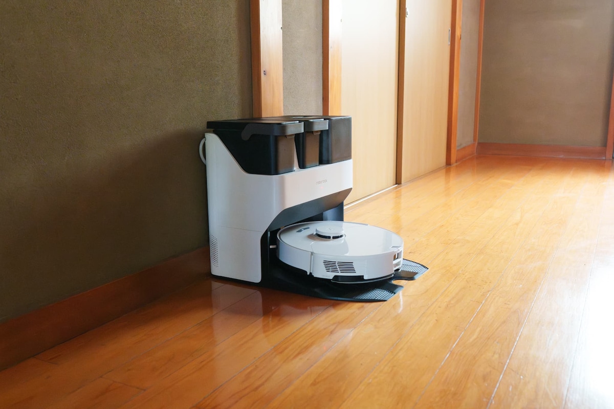 A robotic vacuum cleaner is shown resting on a smooth wooden floor. Natural light highlights the wooden surfaces and the minimalist design of the vacuum. The area appears tidy, with wooden doors leading to other spaces in the background.