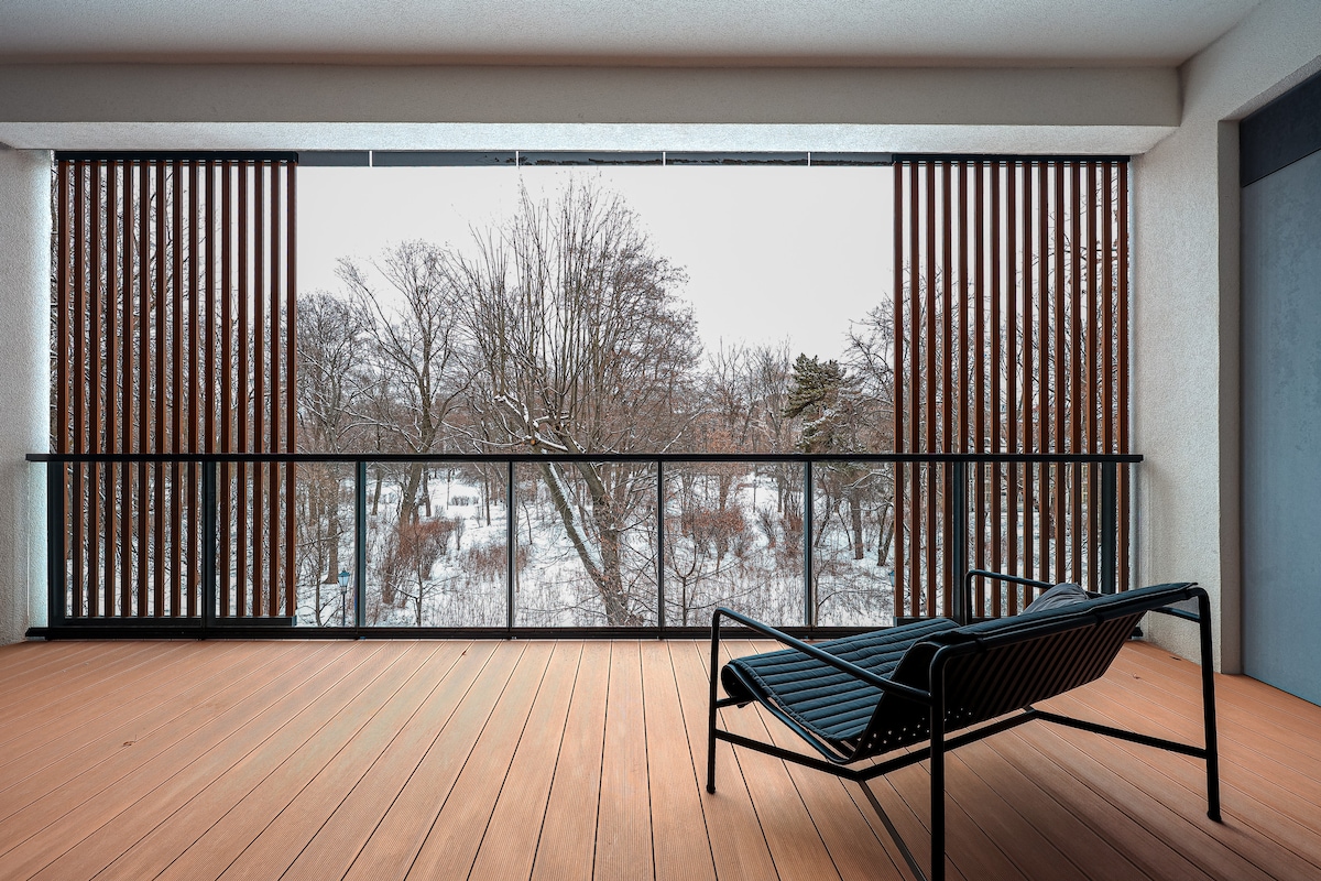 A spacious terrace is featured with wooden flooring and a modern lounge chair, offering views of a snow-covered landscape. Vertical slats provide partial privacy while allowing natural light to filter through. The serene surroundings create a calm atmosphere away from the city.