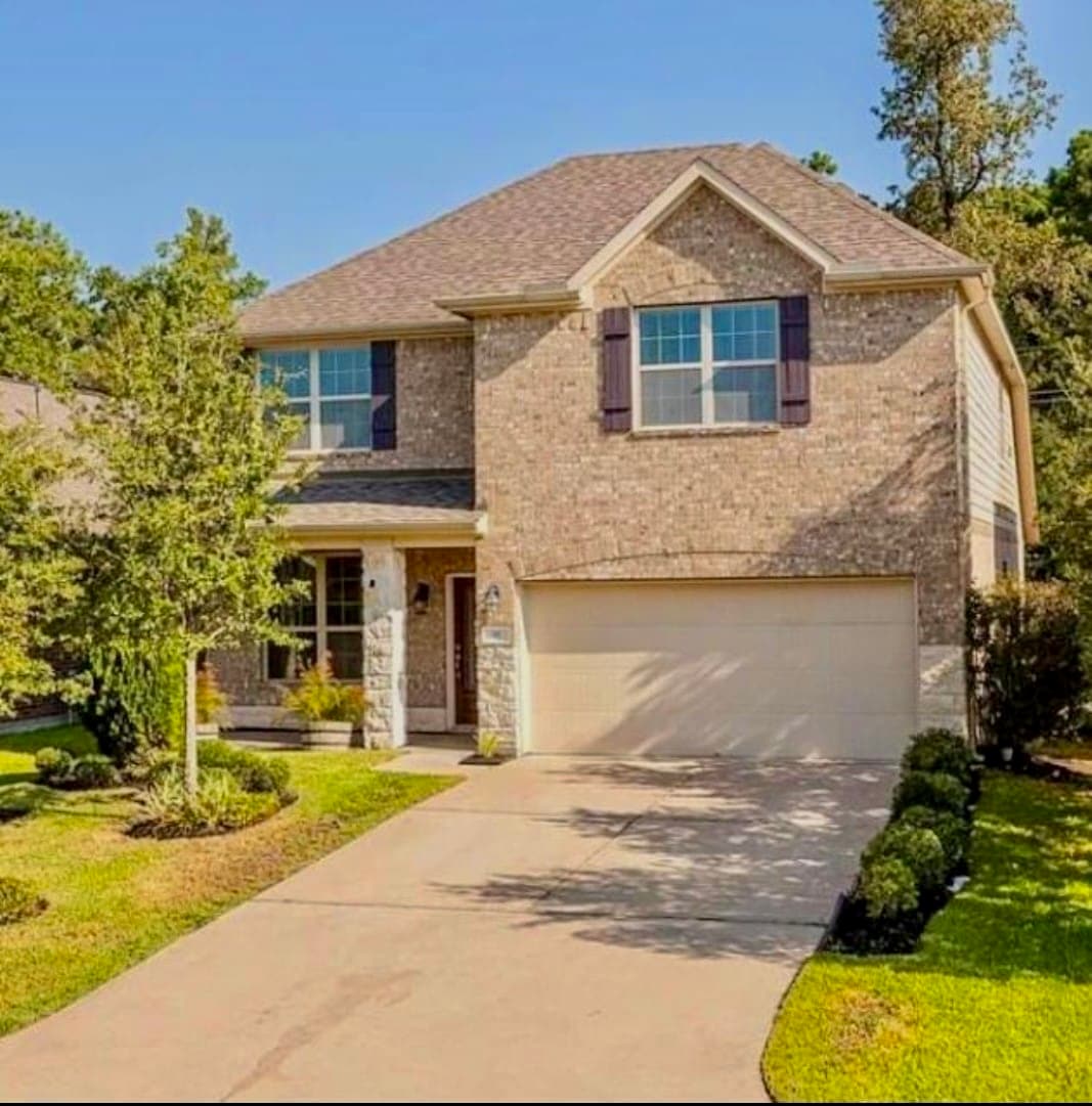 The exterior of the house features a brick facade with two large front windows framed by dark shutters. A neatly paved driveway leads to a garage with a door. Lush green landscaping surrounds the property, enhancing the inviting atmosphere.