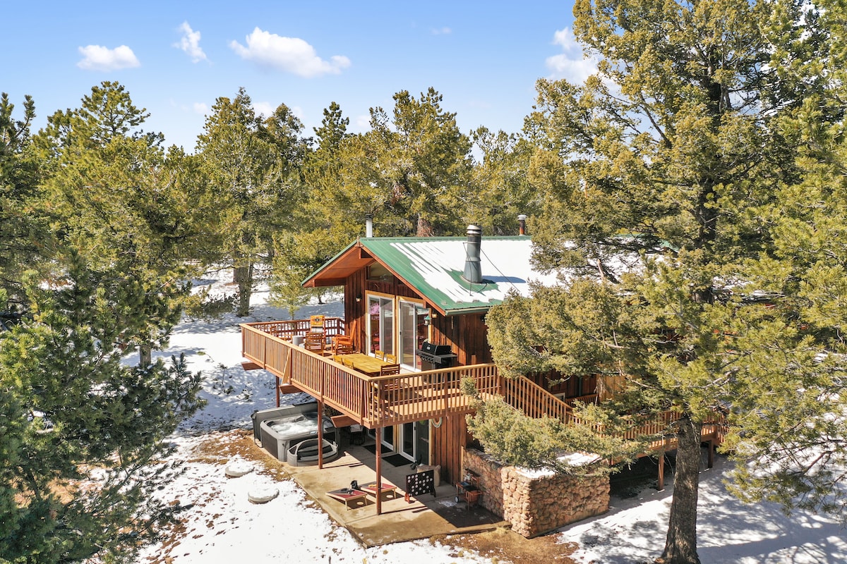 The cabin is nestled among tall pine trees with a snow-covered landscape surrounding it. A wooden deck extends from the structure, featuring a private hot tub and seating areas. The sloped roof and warm wood siding complement the natural setting under a clear blue sky.