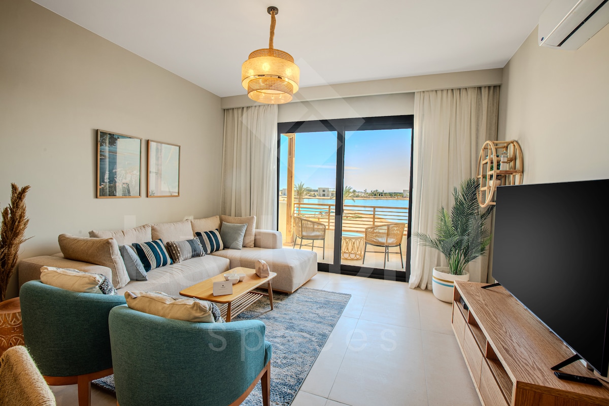 A cozy living area is presented with a light-colored sectional sofa and complementary armchairs. Large glass doors invite natural light and offer views of the outside patio area. Decorative accents include wall art and a stylish overhead light fixture, enhancing the modern aesthetic.