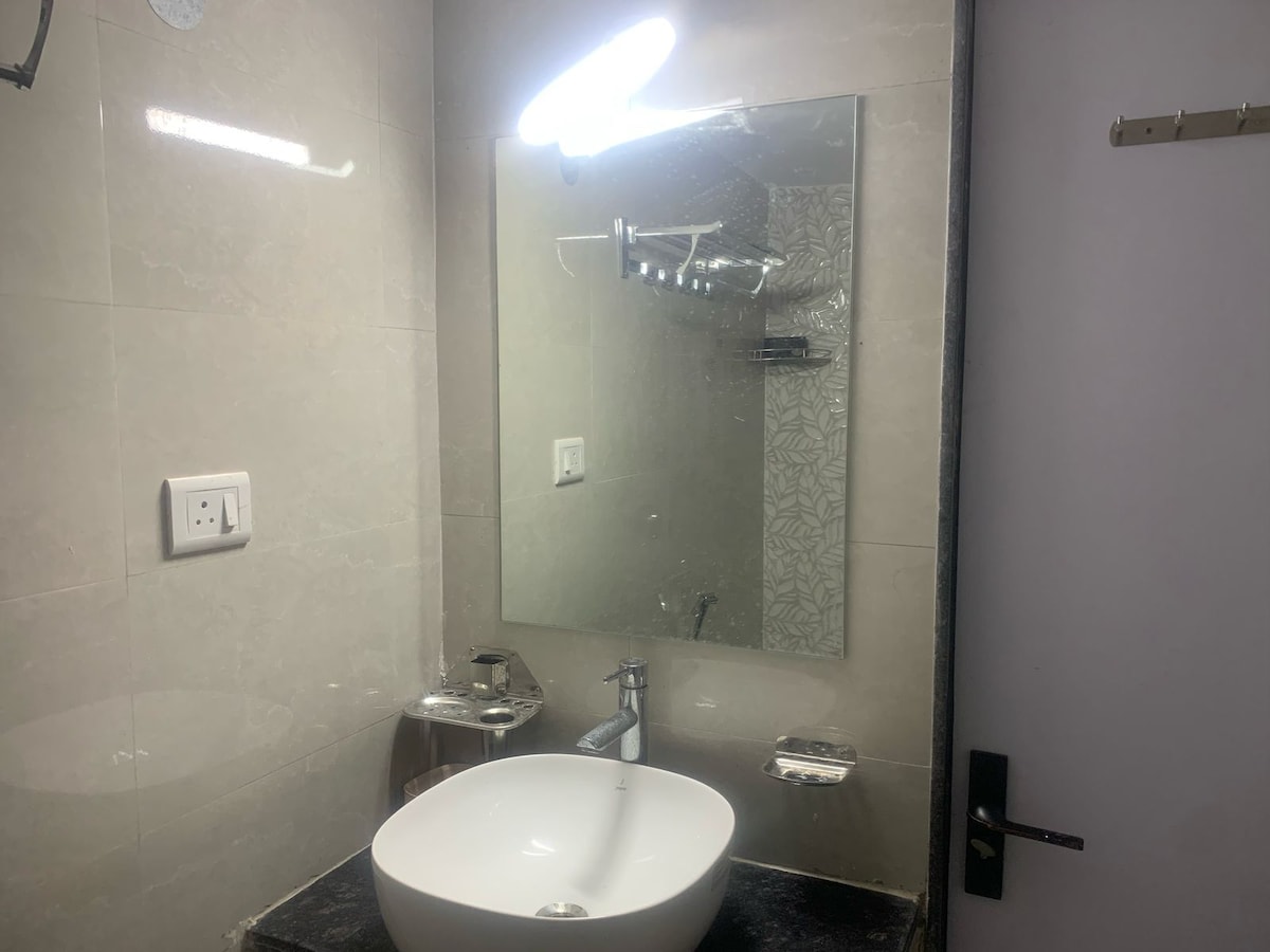 The attached bathroom features a modern design with subtle gray tiles. A circular sink is situated atop a dark countertop, complemented by a large mirror that enhances the room's brightness. Subtle lighting contributes to the clean aesthetic of the space.