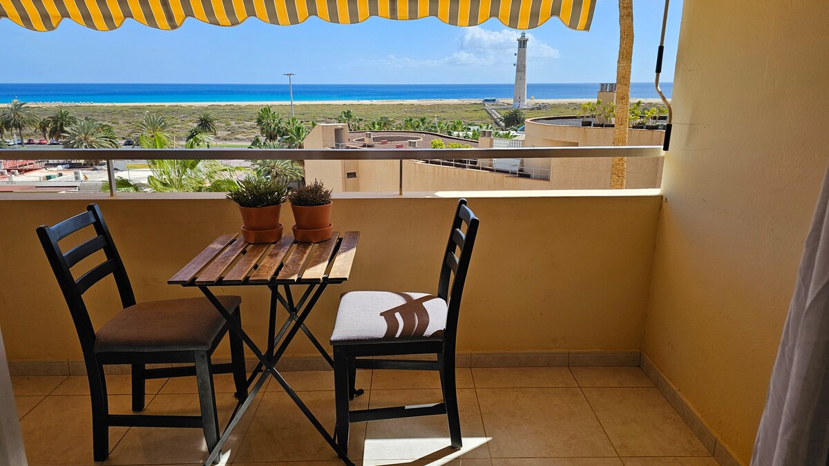 A small outdoor terrace features a wooden table with two chairs, set against a sunny backdrop. Potted plants are placed on the table, while a bright awning provides shade. In the distance, a lighthouse and the coastline are visible, enhancing the tranquil setting.