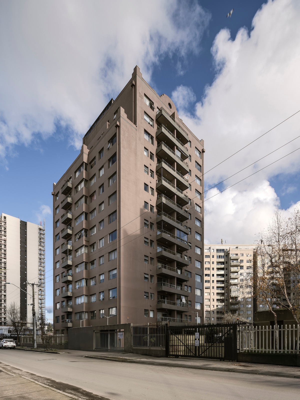 A modern multi-story building is depicted, featuring a sleek exterior in neutral tones. The structure showcases multiple balconies with glass railings. Surrounding buildings and a clear sky can be seen in the background, contributing to the urban environment.