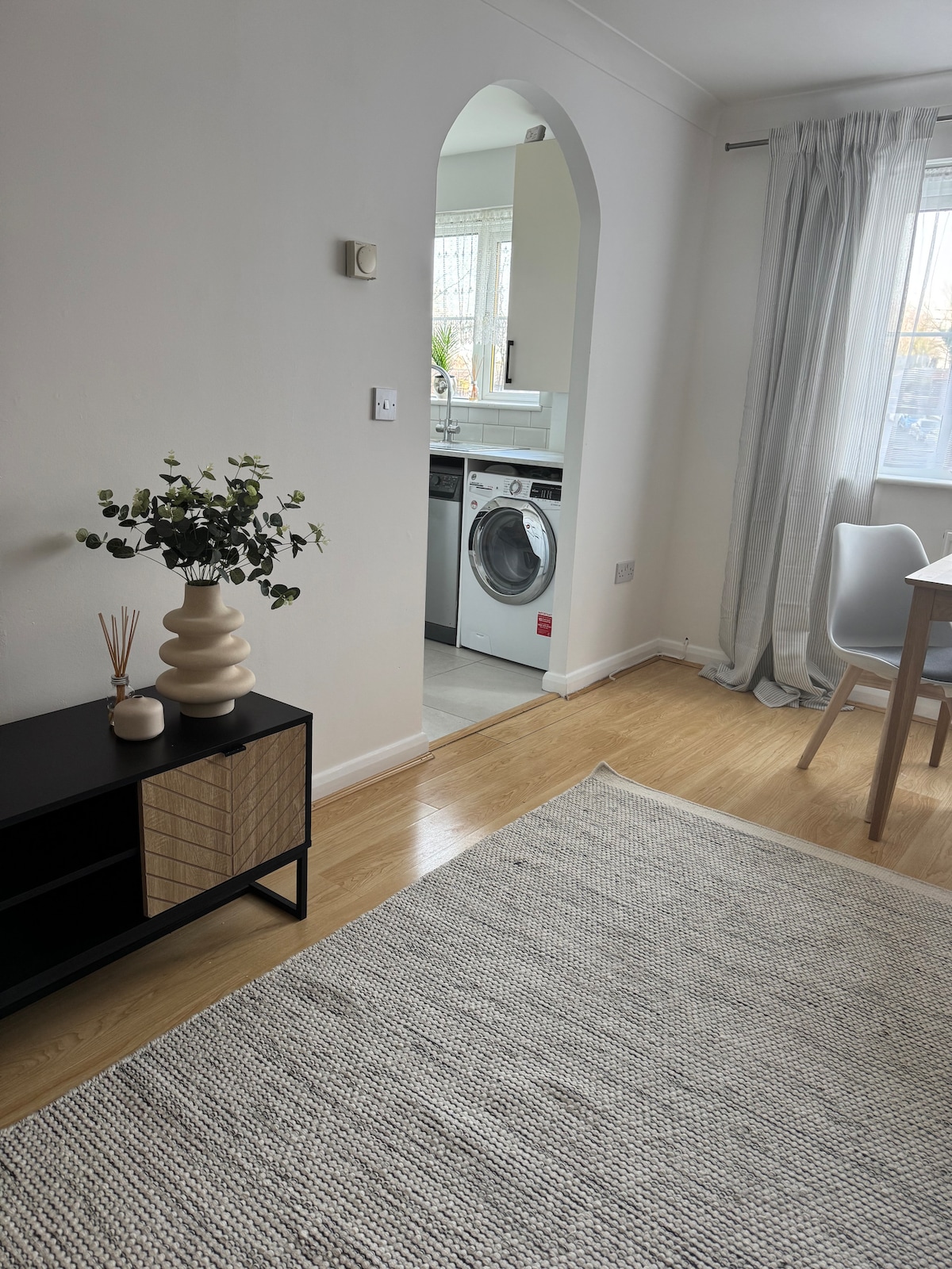 A clean and modern living area is visible, featuring a textured rug on light laminate flooring. An elegant plant arrangement is positioned on a low cabinet. An archway leads to a bright kitchen area where a washing machine is located, with natural light streaming through a nearby window.