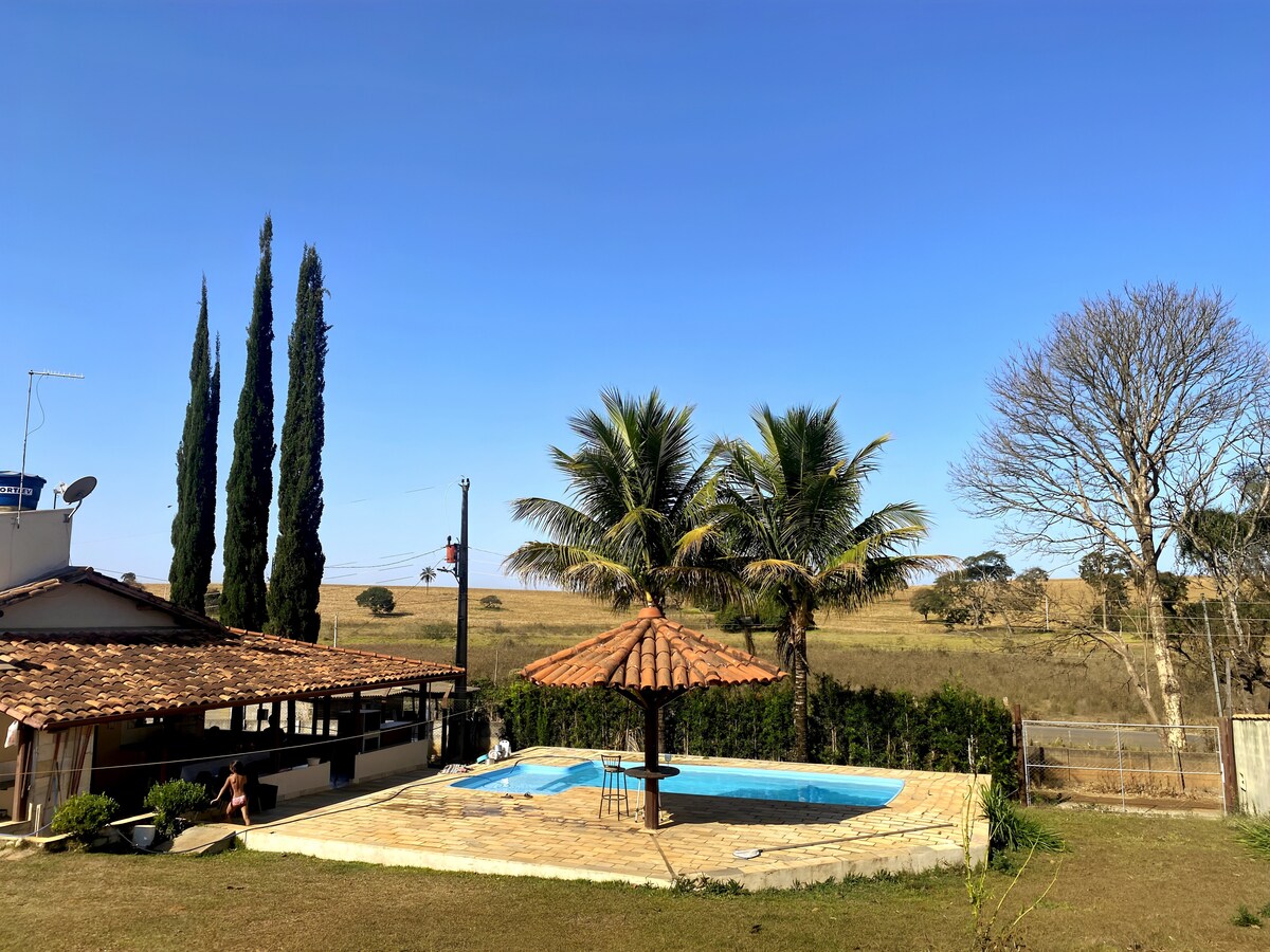A tranquil outdoor area showcases a swimming pool surrounded by a stone deck and palm trees, with a thatched-roof gazebo providing shade. In the background, open fields and a clear blue sky enhance the serene atmosphere.