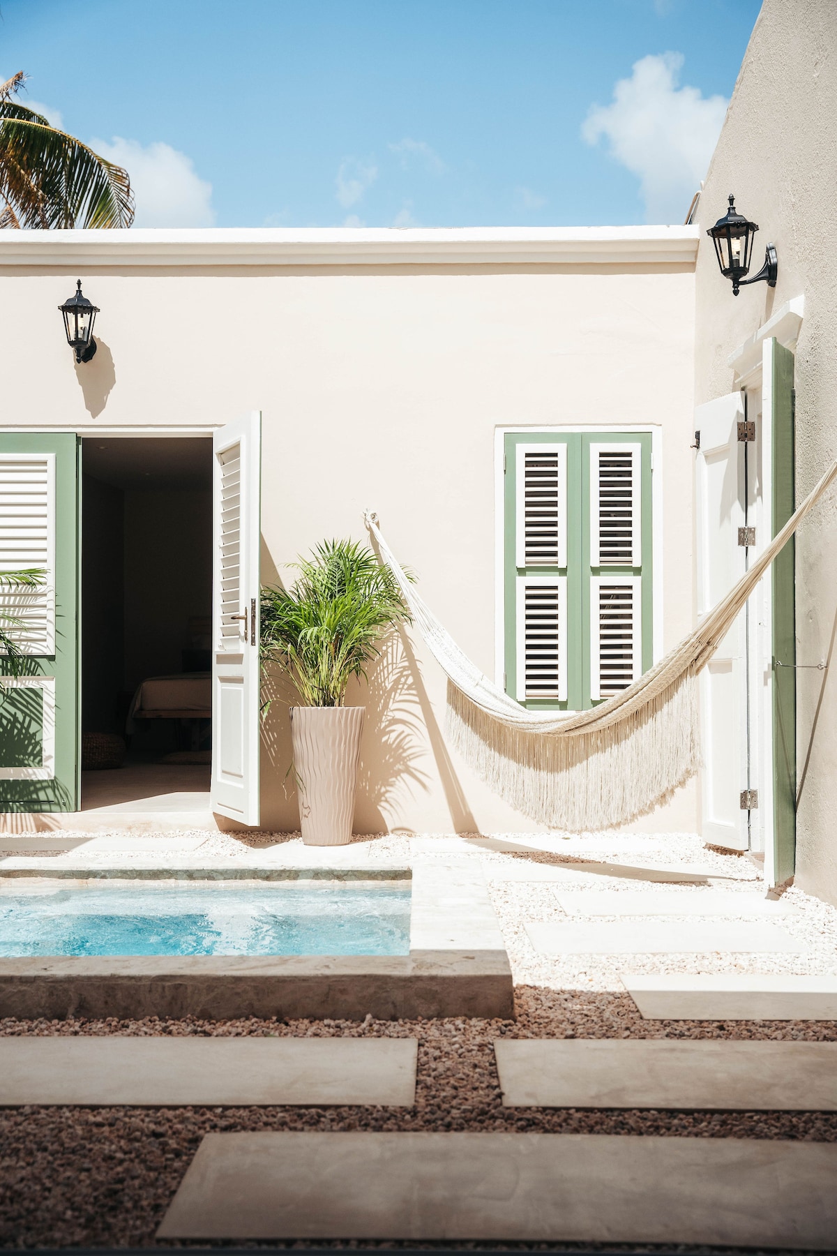 A private courtyard showcases a serene plunge pool with clear water, surrounded by natural stone flooring. Light-coloured walls frame open doors revealing an interior space. A hammock hangs gracefully between two charming green-shuttered windows, complemented by a tall potted plant adding a touch of greenery.