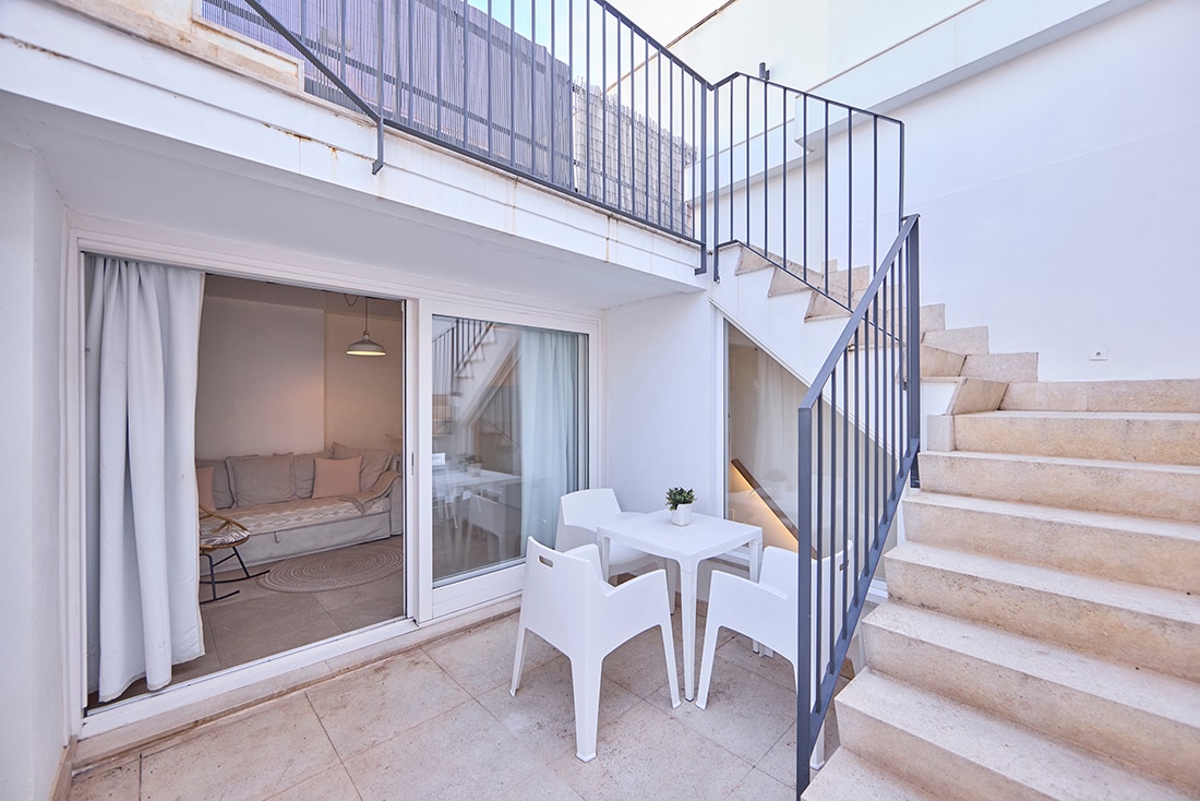 A small outdoor seating area is visible, featuring a white table and two matching chairs. A set of stairs leads to an upper level, while the entry to the living space is framed by large glass doors, revealing a cozy interior.