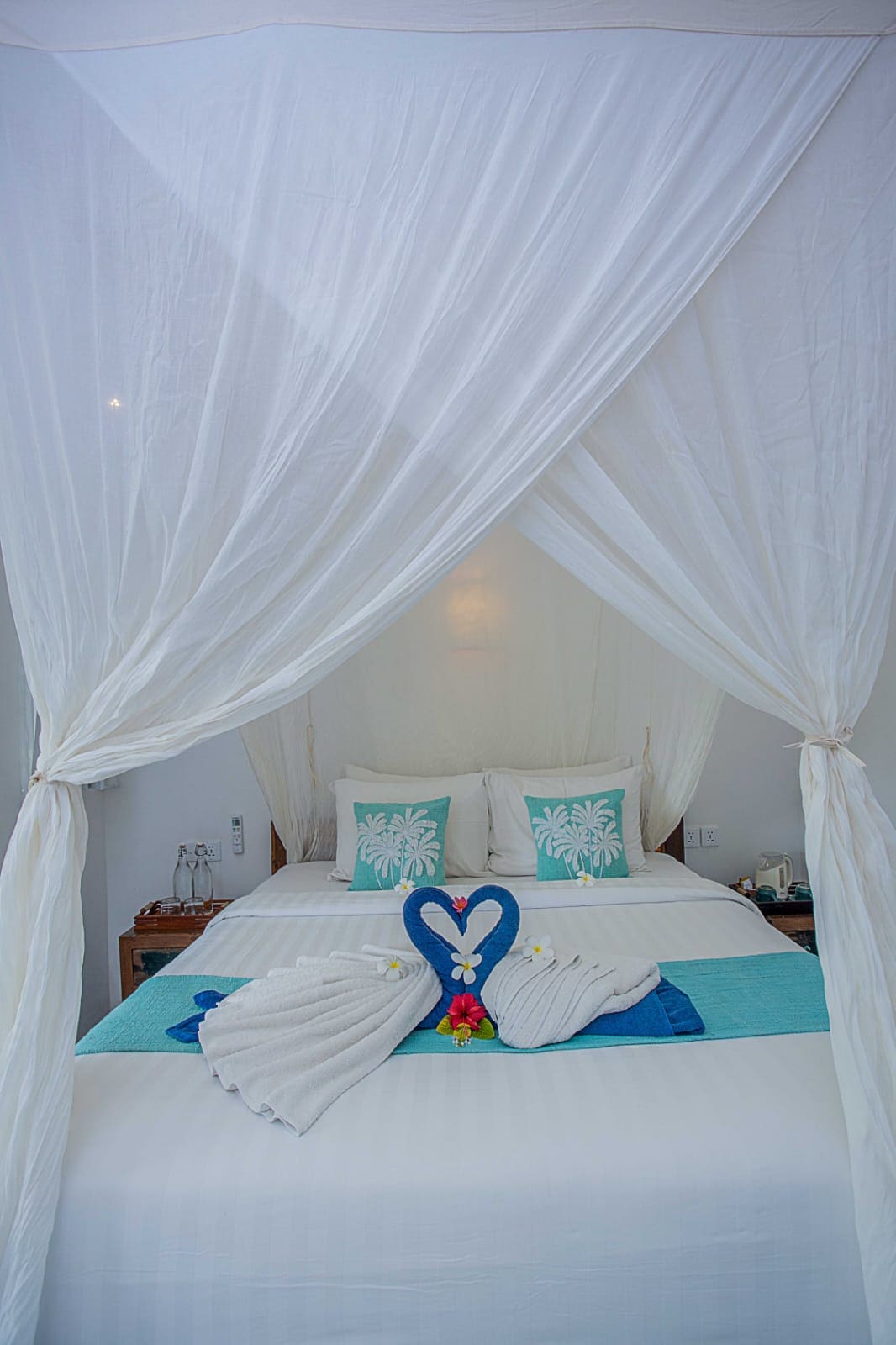 A serene bedroom is framed by sheer white drapery, creating a cozy atmosphere. The spacious bed features turquoise accents and decorative pillows with palm motifs. A heart-shaped towel display and floral touches add a welcoming detail, enhancing the room's tranquil ambiance.