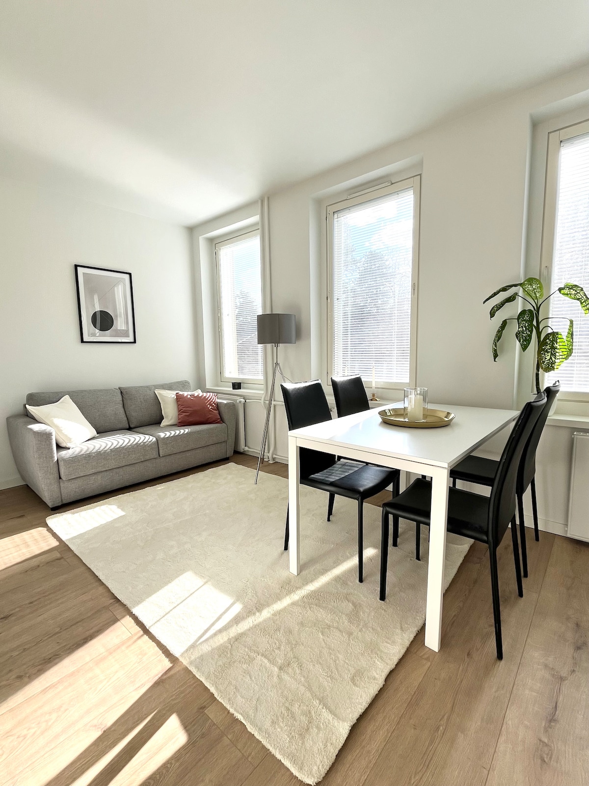 A well-lit living area features a light-colored area rug and a gray sofa adorned with a decorative pillow. A white dining table surrounded by four black chairs is complemented by a green plant in the corner. Sunlight enters through the large windows.