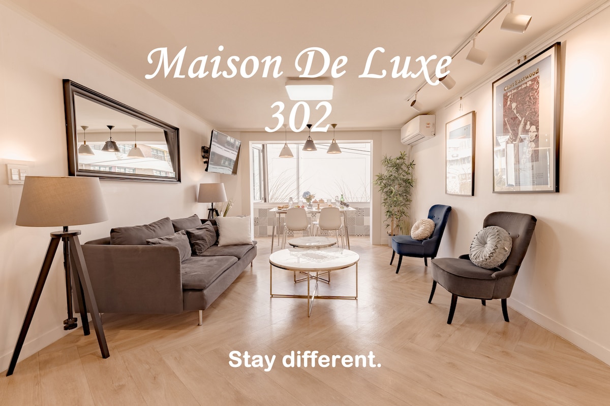 The living area showcases contemporary furniture with a full-sized sofa and accent chairs arranged around a circular coffee table. Natural light floods in through expansive windows, providing a view of the balcony. Elegant lighting fixtures hang from the ceiling, enhancing the modern aesthetic.