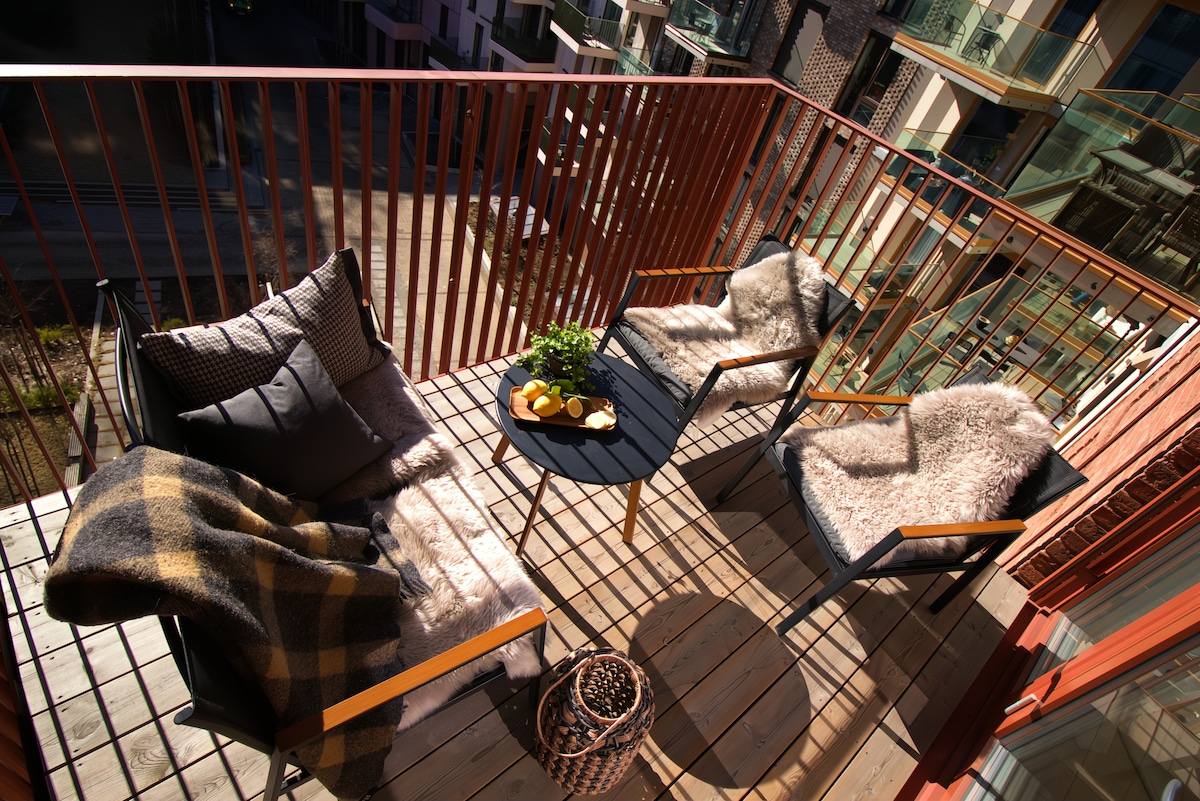 A spacious balcony features two comfortable chairs draped with soft throws, alongside a small table set with fresh fruit. The wooden floor and railing cast structured shadows, enhancing the inviting outdoor seating area.