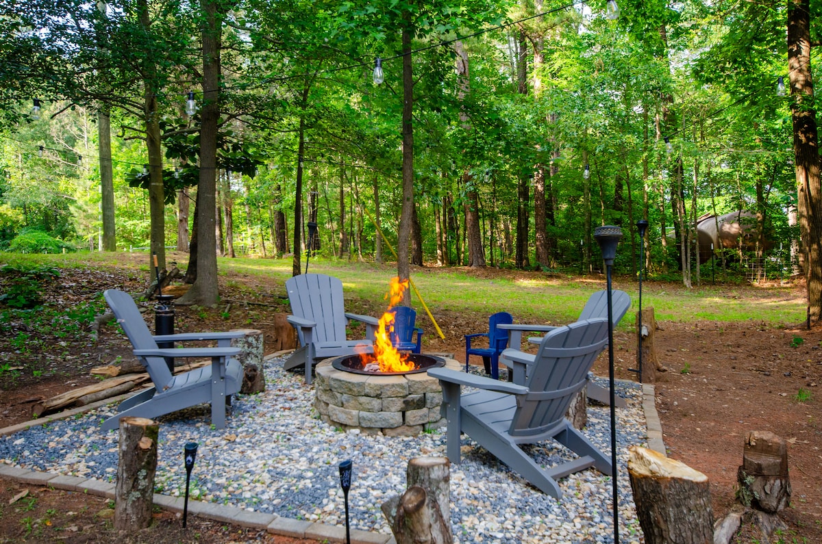 A stone fire pit is surrounded by several adirondack chairs, set against a backdrop of tall trees and natural greenery. The flames flicker warmly, inviting relaxation in a peaceful outdoor space. Pebbles line the ground, enhancing the rustic atmosphere.