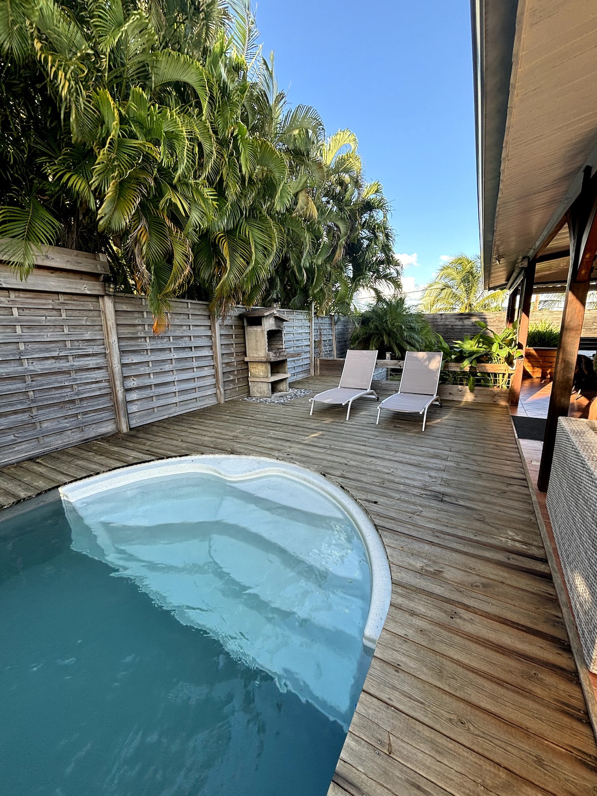A private pool is surrounded by a wooden deck, featuring two lounge chairs positioned for relaxation. Lush green foliage is visible in the background, providing a serene ambiance. A built-in barbecue area is seen to the side, enhancing the outdoor space.