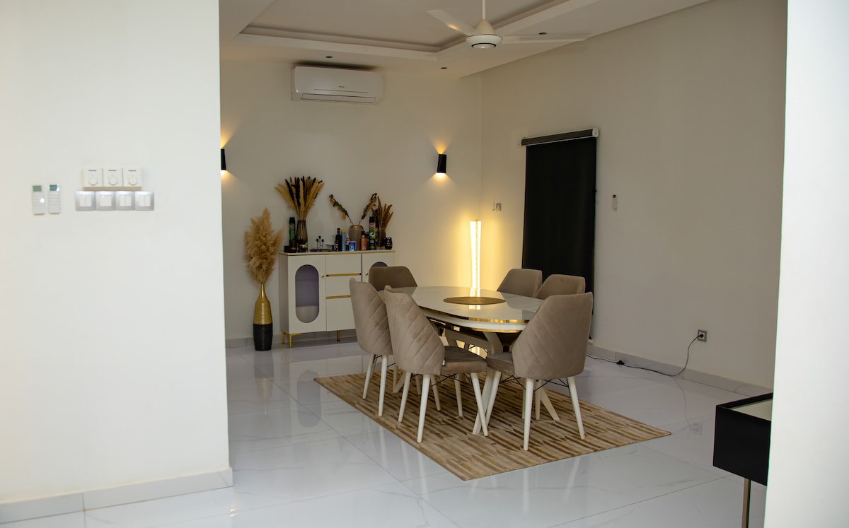 A modern dining area features a round table surrounded by six cushioned chairs. A textured area rug lies beneath the table, while a sideboard displays decorative elements. Soft lighting is provided by floor lamps and wall sconces, contributing to a calm environment.